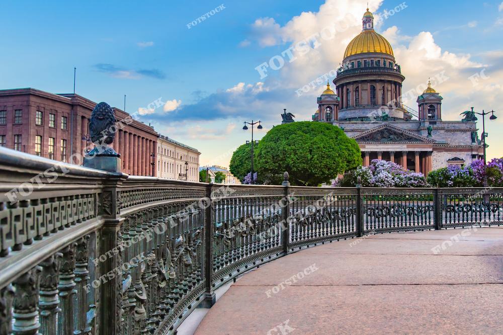 На фото изображён Исаакиевский собор — один из главных архитектурных символов Санкт-Петербурга. На переднем плане — изящное литое ограждение и аллея, украшенная цветущими сиреневыми кустами. Справа видна часть Исаакиевской площади, а слева — исторические здания Адмиралтейского района. Золотой купол собора ярко выделяется на фоне голубого неба. Картина передаёт атмосферу спокойного весеннего утра в центре города.